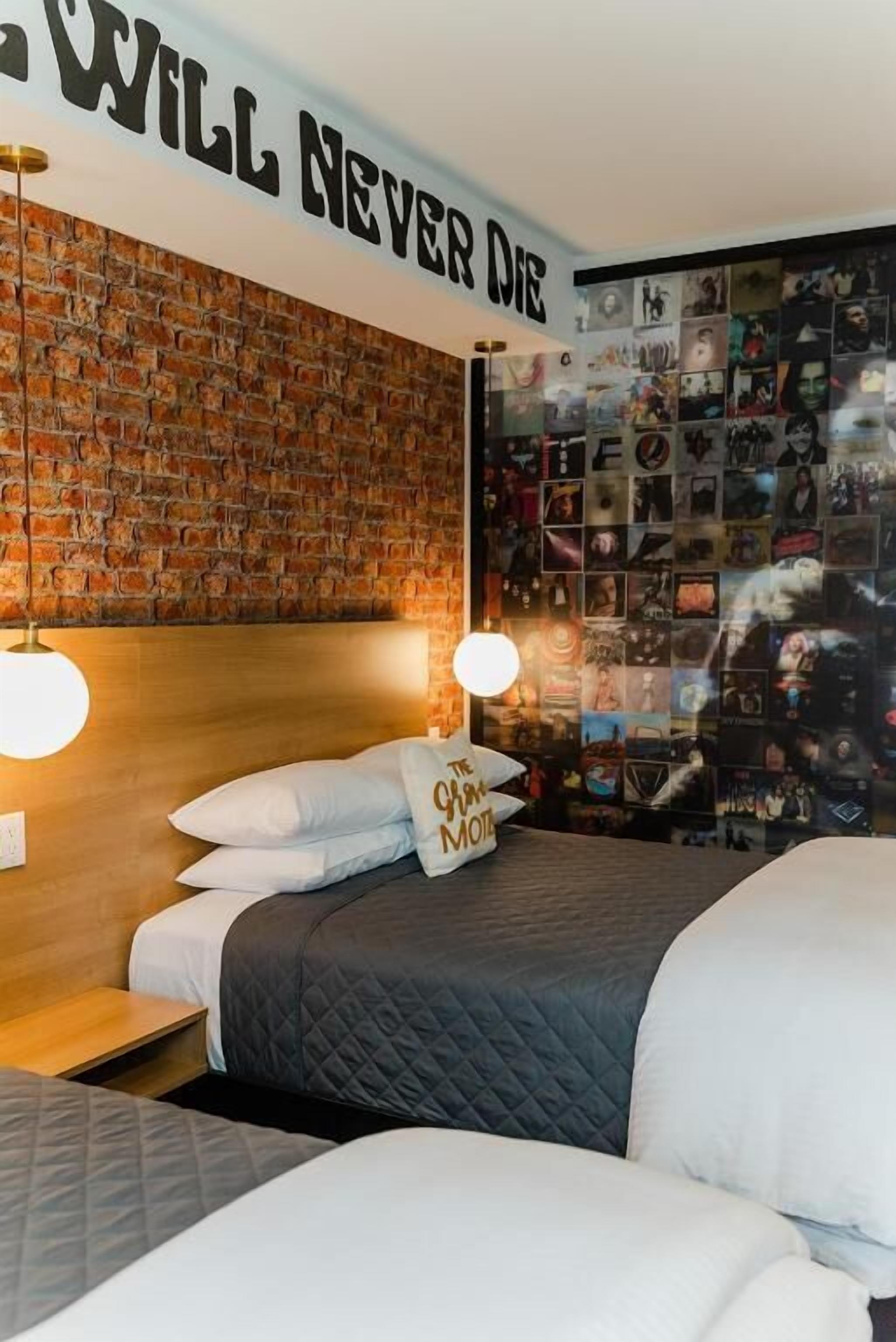 Um quarto com parede de tijolos, uma cama com roupa de cama branca e cinza, uma parede com a placa 'WILL NEVER DIE' e uma colagem de fotografias na parede.