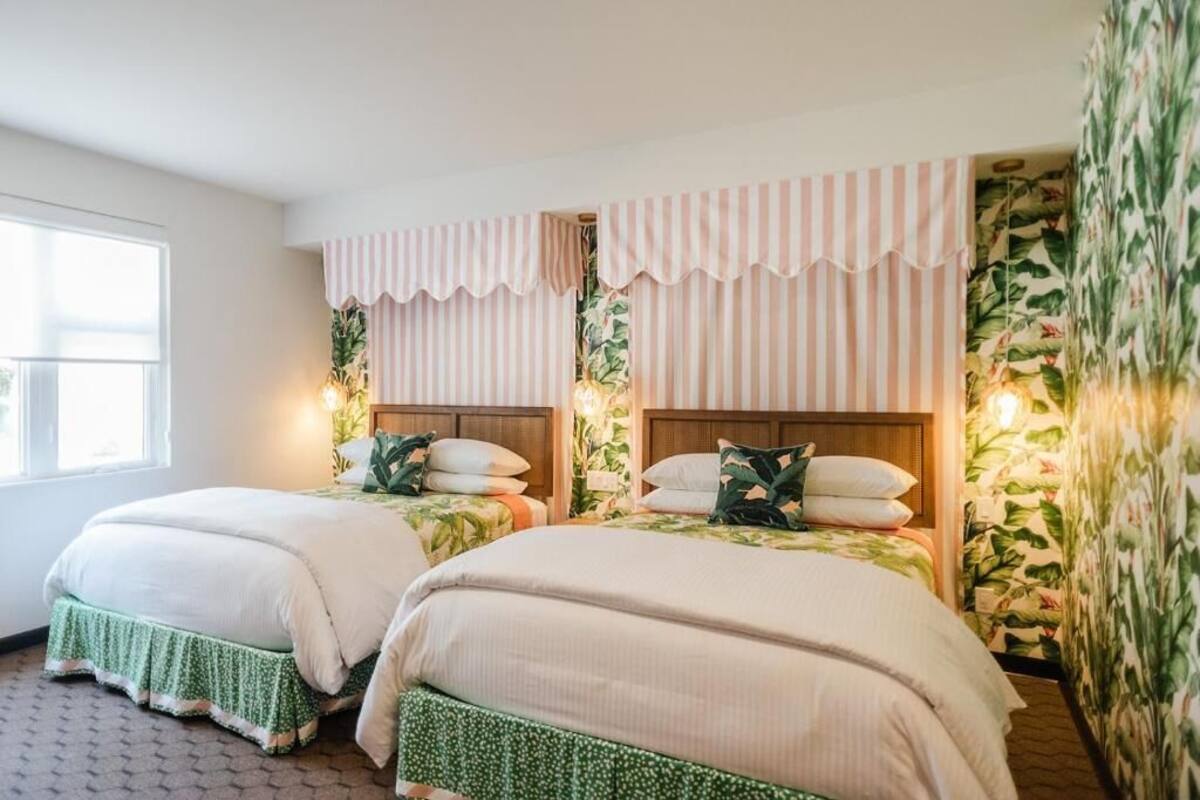 A room with two beds, a window, and a wall with a tropical wallpaper.