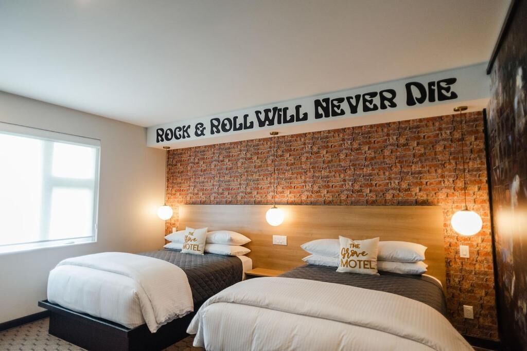 A hotel room with two beds, a brick wall, and a sign that reads 'ROCK & ROLL WILL NEVER DIE'.