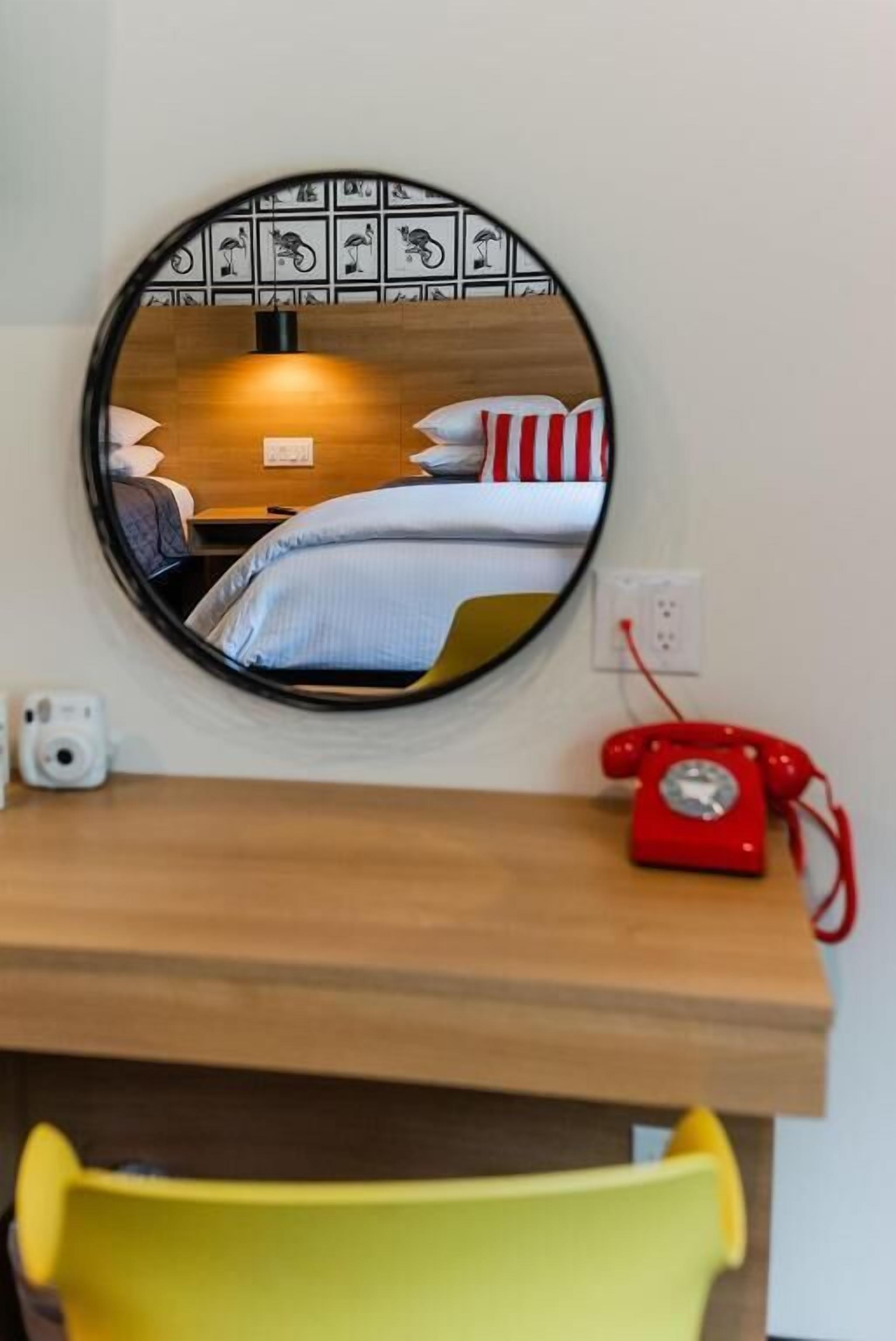 Un miroir rond reflète un lit avec une literie à rayures, une tête de lit en bois, un téléphone rouge et un mur orné de carreaux décoratifs.