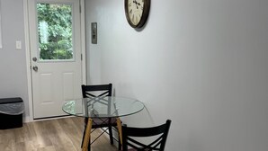 Un comedor con una mesa redonda de cristal, dos sillas, un reloj de pared y una puerta que da a zonas verdes.
