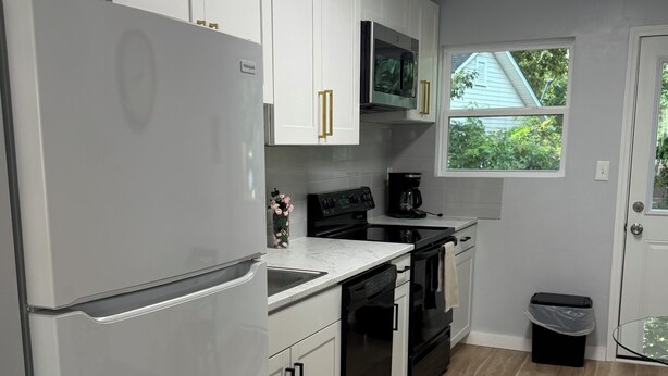 Una cocina moderna con armarios blancos, un refrigerador, un microondas, una cafetera y una ventana con vistas a zonas verdes.