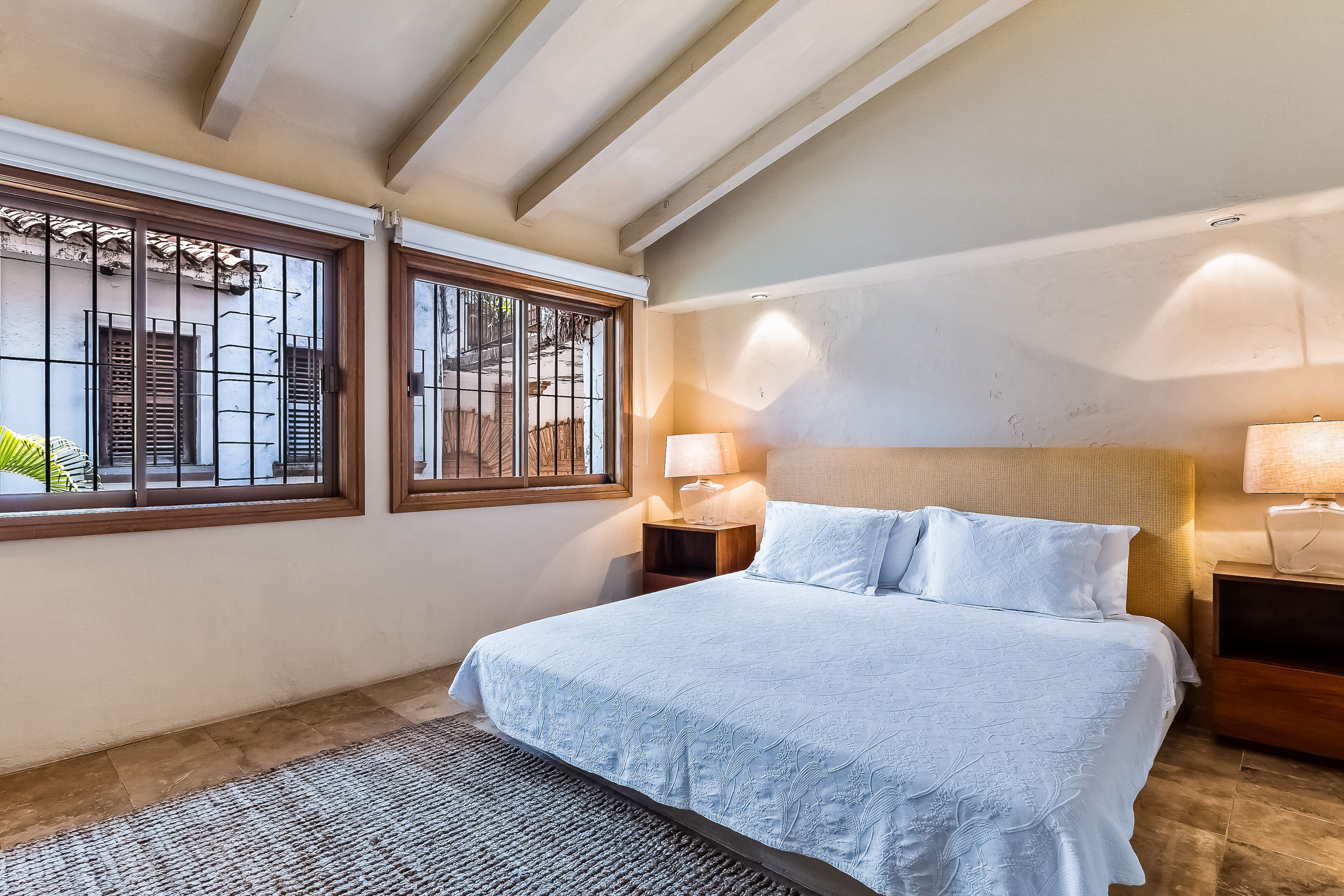 Un dormitorio con una cama, dos mesitas de noche con lámparas, un ventanal con barrotes y un techo con vigas a la vista.