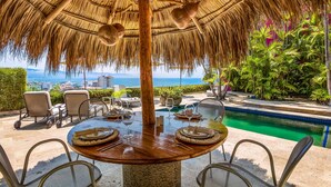 A round table set for dining with a thatched roof overhead, surrounded by chairs, and overlooking a swimming pool with a view of the ocean and cityscape. - Spectacular Hillside Villa With Pool, Incredible Views (Puerto Vallarta)