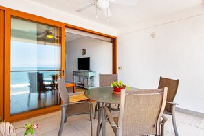 A balcony with a glass door leading to a dining area with a table and chairs, a ceiling fan, and a television in the background. - Beachfront 4th-floor Condo With Views, Outdoor Pool, Balcony, W/D & Central AC (Nuevo Nayarit)