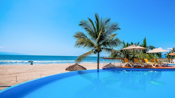 Ein Pool mit Blick auf Strand und Meer, Liegestühle und Palmen.