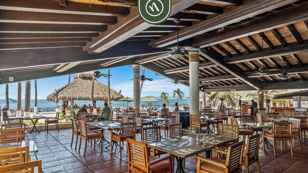 Ein Restaurant im Freien mit Holztischen und -stühlen, einem Reetdach und Blick auf das Meer.