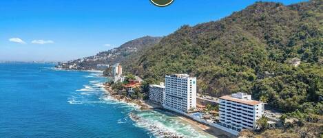 A coastal hotel with multiple floors, located near a rocky shoreline and a forested hillside.