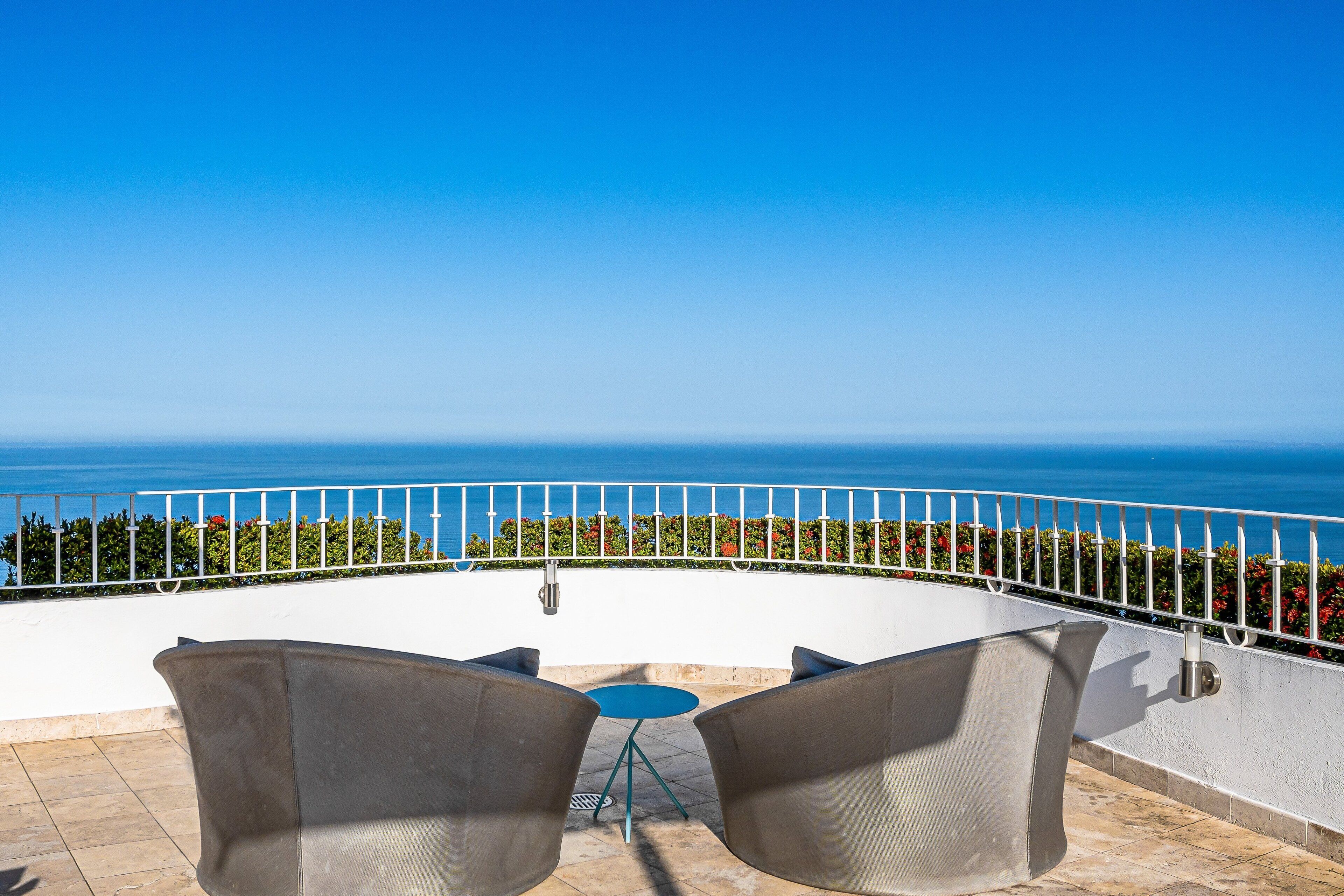 A terrace with a view of the sea, metal chairs, a small round table, and a white railing.