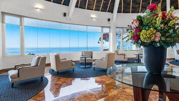Un hall d’entrée spacieux, doté d’une grande baie vitrée donnant sur la mer, d’un coin salon avec des fauteuils et une table, ainsi que d’un grand vase rempli de fleurs.