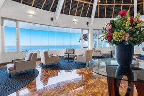 A spacious lobby with a large glass window offering a view of the sea, a seating area with chairs and a table, and a large vase of flowers. - Upscale Bayview Villa With hot Tub, Private Terrace, Pool & Stylish Interiors (Puerto Vallarta)