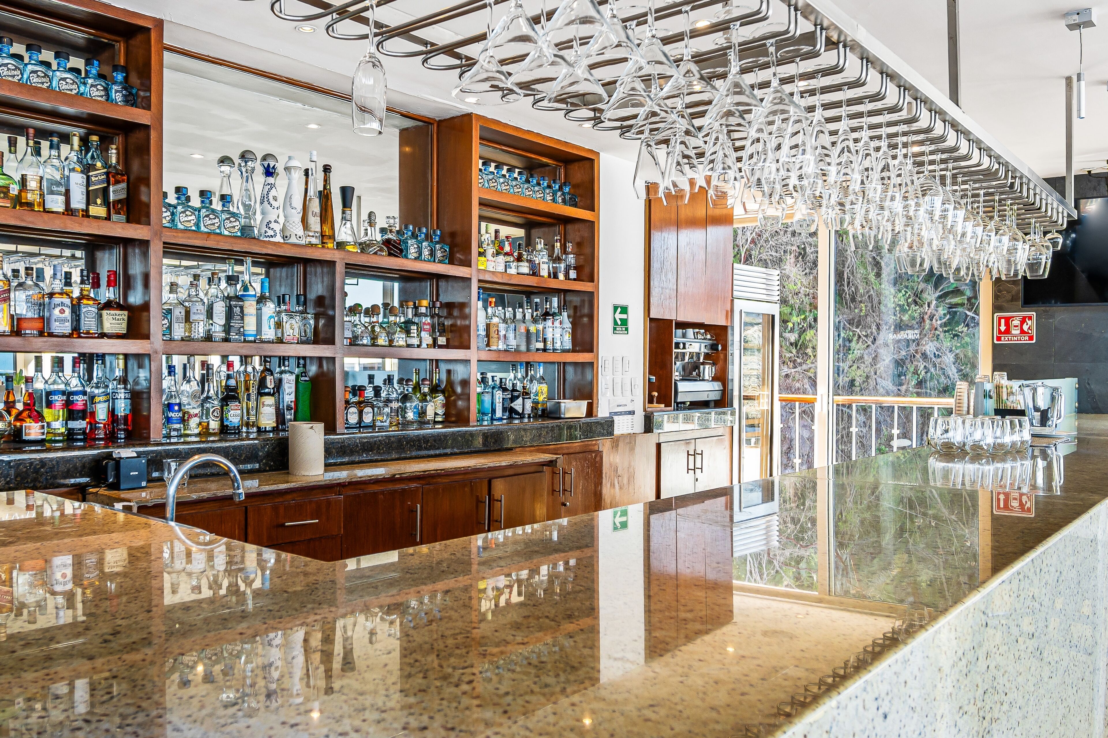 Un espace bar avec des étagères remplies de bouteilles d’alcool, un comptoir en granit poli et un luminaire suspendu.