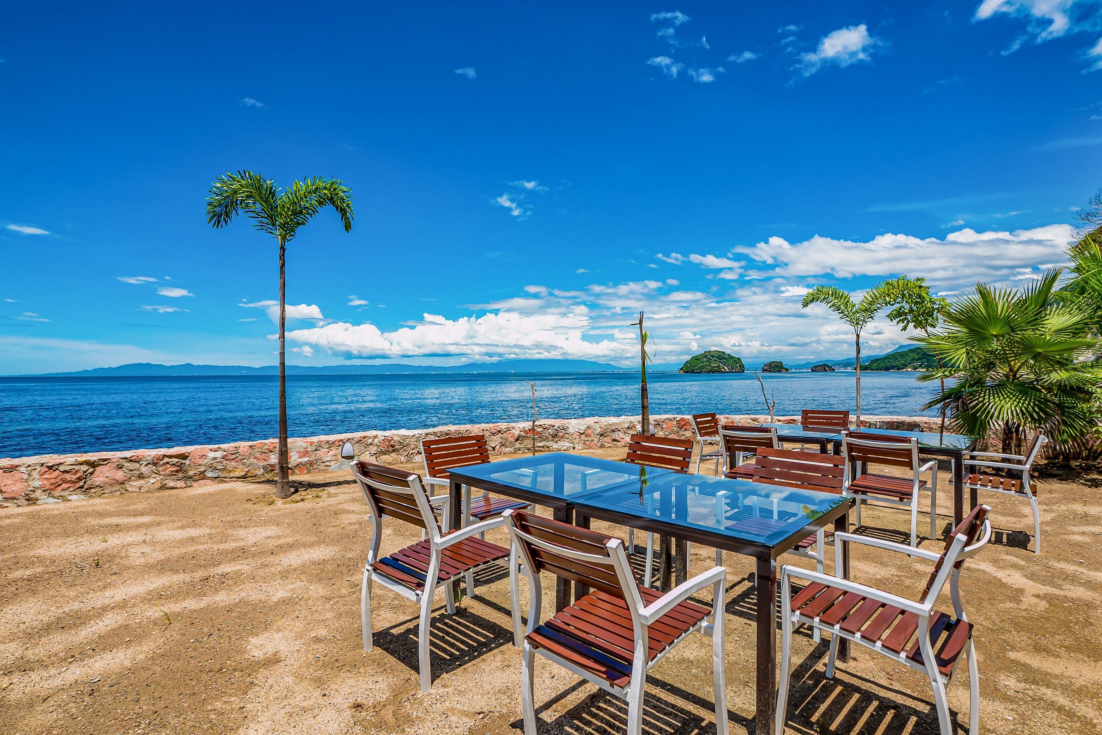 A beachside dining area with a glass table and chairs, palm trees, and a view of the sea.