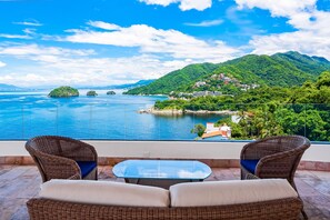 Varanda com mobiliário de vime e mesa de vidro, com vista para uma paisagem costeira com montanhas e um corpo de água.