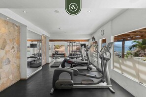 A modern gym with treadmills, ellipticals, and weight machines, featuring a stone wall and large windows with a view of the outdoors. - Peaceful Oceanview Studio - Costa Sur Estudio 10 (Puerto Vallarta)