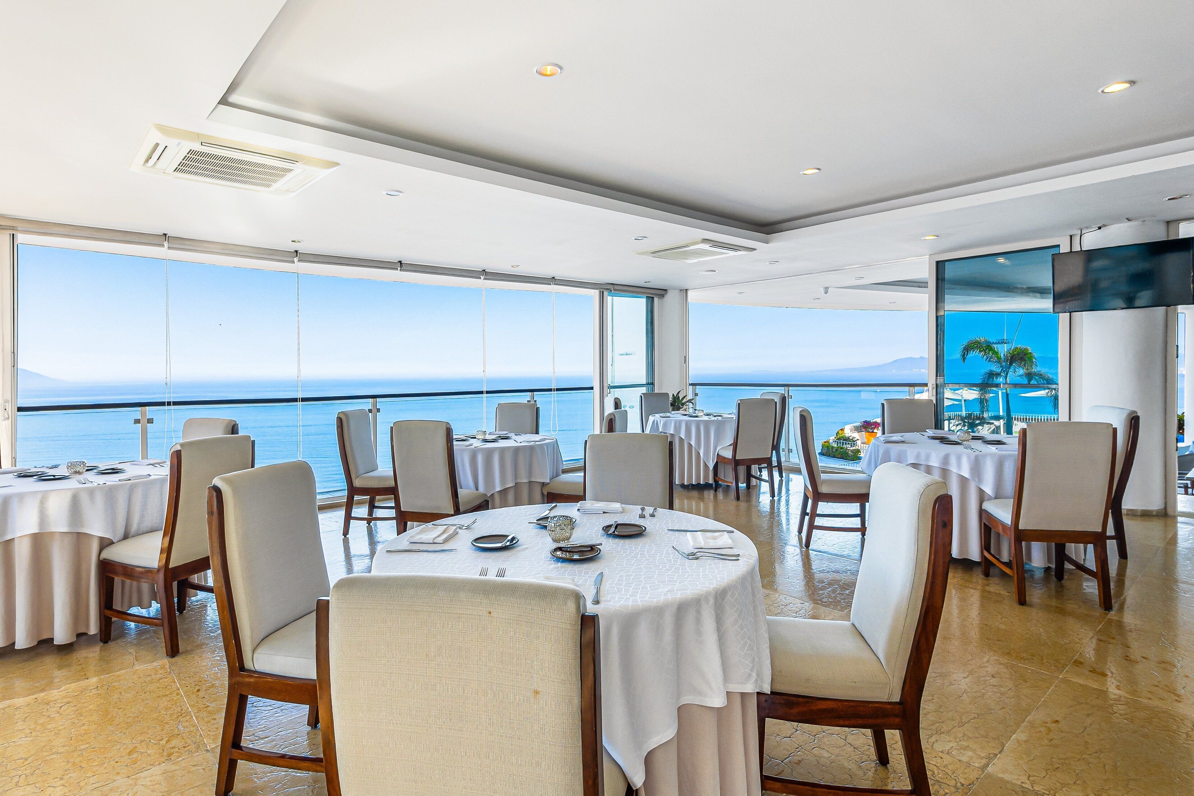 A dining area with round tables, chairs, and a large window with a view of the sea.
