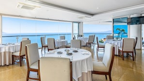 A dining area with round tables, chairs, and a large window with a view of the sea. - Elegant Hillside Villa With Panoramic Views, Pool, hot tub & Luxe Interiors (Puerto Vallarta)