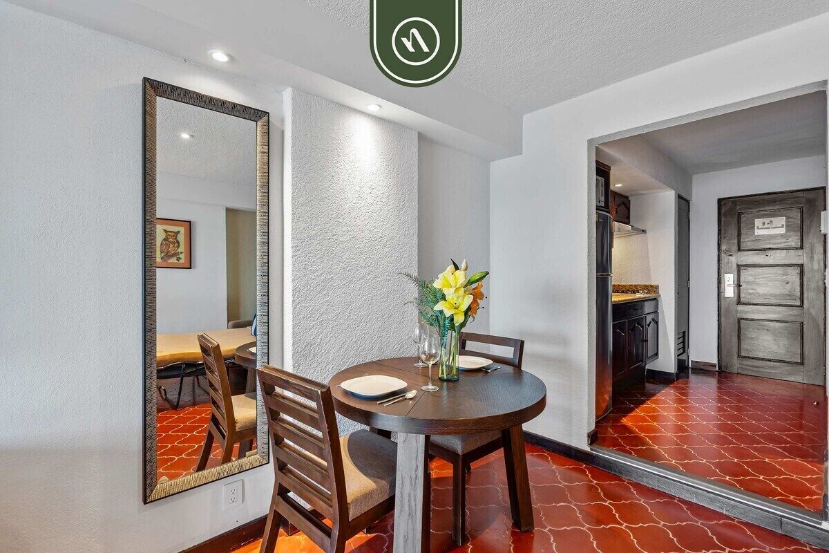 Un comedor con una mesa redonda, sillas, un jarrón con flores, un espejo grande y una puerta que da a otra habitación.