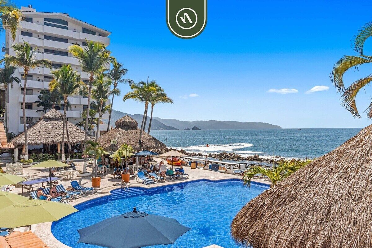 A pool area with lounge chairs and umbrellas, a building with balconies, and a view of the ocean.