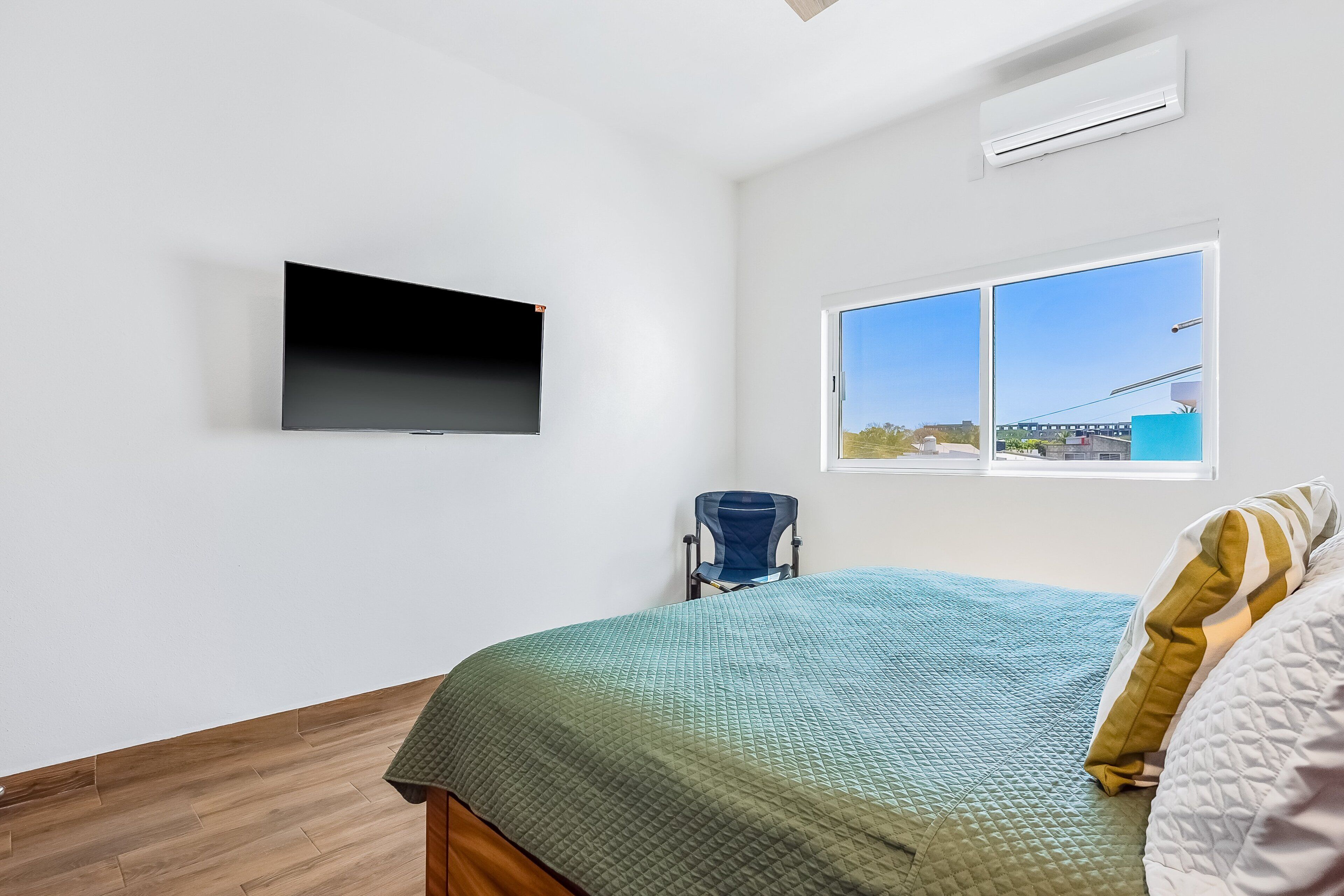 A bedroom with a bed, a TV mounted on the wall, a window with a view, and an air conditioning unit.