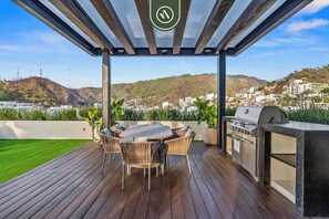 Eine Terrasse im Freien mit Holzdeck, einem runden Tisch mit Korbstühlen, einem Grill und Blick auf eine Hügelstadt.
