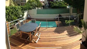 Una terraza de madera con mesa y sillas, una piscina y una valla verde.