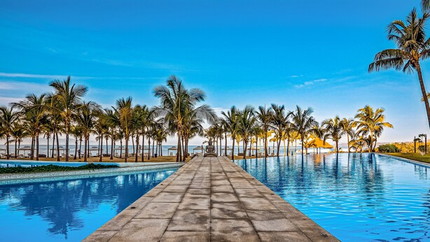 Ein langer Pool mit gefliestem Gehweg, umgeben von Palmen und blauem Himmel.
