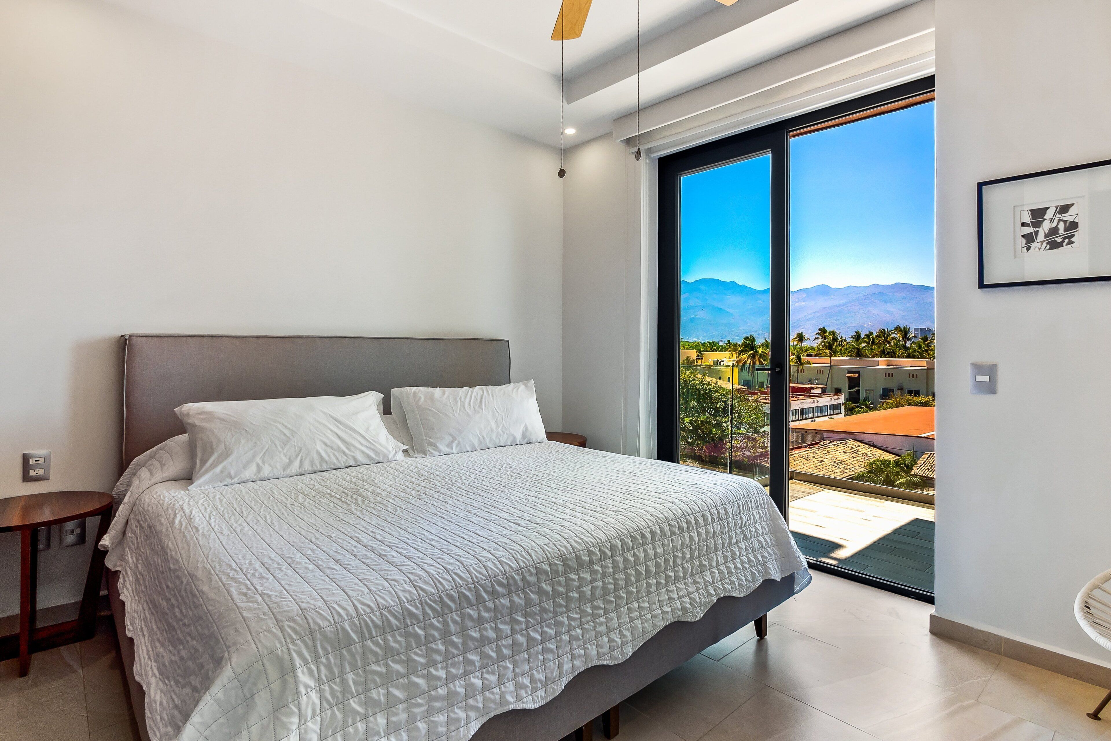 Une chambre à coucher comprenant un lit, une table de chevet, une grande fenêtre donnant sur des immeubles et des montagnes, et un ventilateur de plafond.