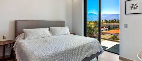 A bedroom with a bed, a bedside table, a large window with a view of buildings and mountains, and a ceiling fan.