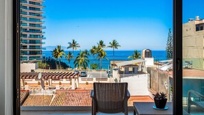 A balcony with a view of the ocean, palm trees, and a building. - 5th-floor Condo With Partial Ocean Views, Heated Pool, Sauna, Gym, Balcony, W/D (Puerto Vallarta)