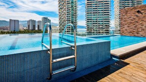 A rooftop swimming pool with a metal ladder, surrounded by high-rise buildings and a clear sky. - 5th-floor Condo With Partial Ocean Views, Heated Pool, Sauna, Gym, Balcony, W/D (Puerto Vallarta)