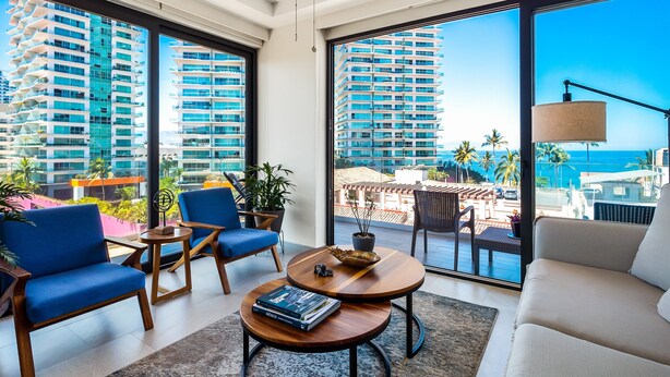 A modern living room with large windows, a ceiling fan, and a view of tall buildings and the ocean. - 5th-floor Condo With Partial Ocean Views, Heated Pool, Sauna, Gym, Balcony, W/D (Puerto Vallarta)