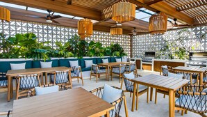 An outdoor dining area with wooden tables and chairs, a grill, and a patterned wall. - Modern Condo With Rooftop Pool, Balcony, gas Grill & gym Access (Puerto Vallarta)