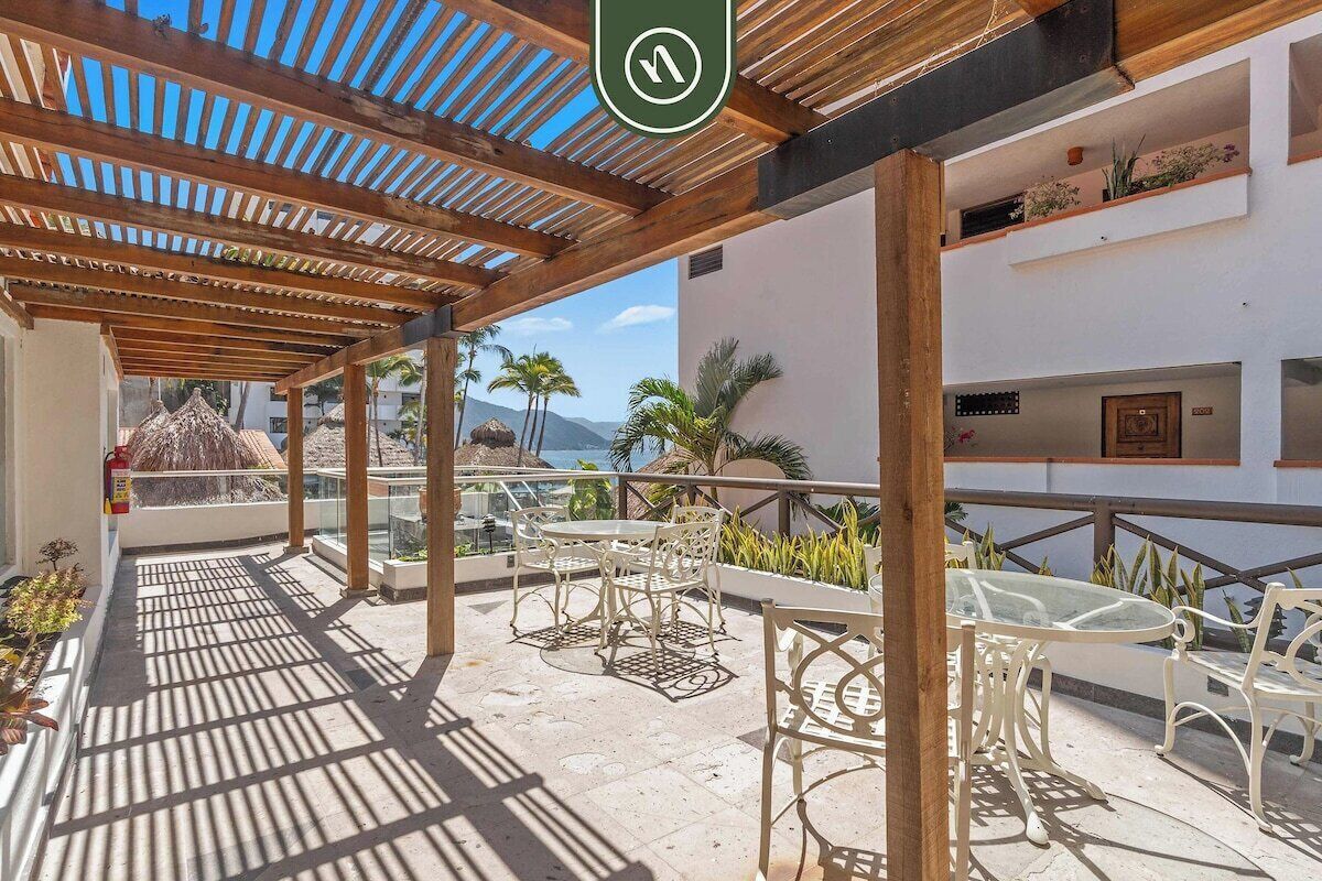 Ein überdachter Außenbereich mit einer Holz-Pergola, weißen Metallmöbeln und Blick auf Meer und Berge im Hintergrund.