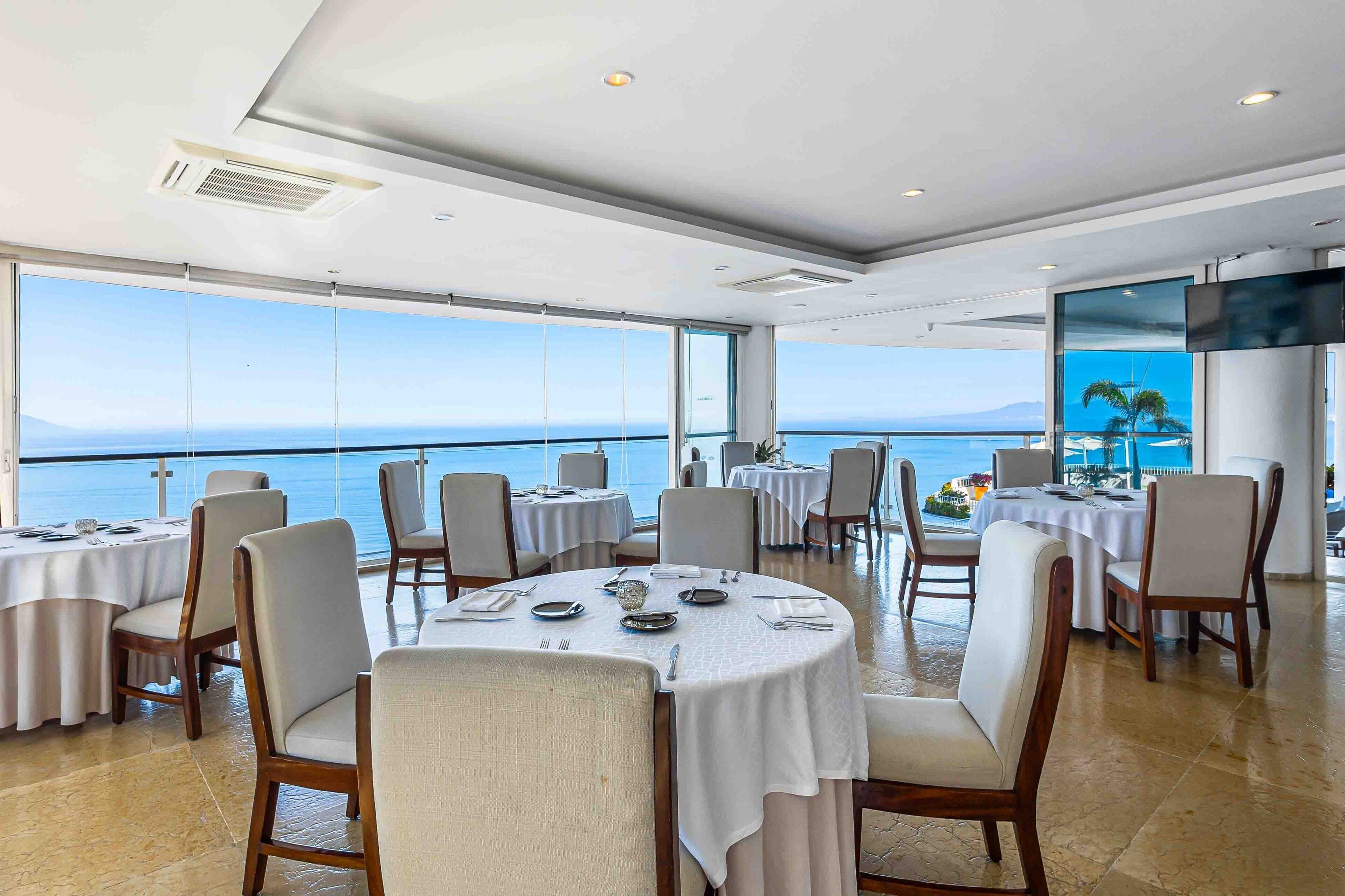 A dining area with round tables, chairs, and a large window overlooking the sea.