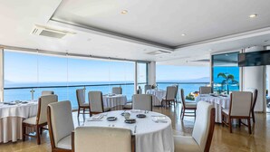 Een eetgedeelte met ronde tafels, stoelen en een groot raam met uitzicht op de zee.