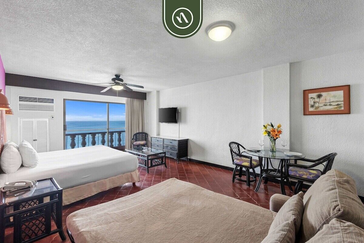 A hotel room with a bed, a dining table with chairs, a ceiling fan, a TV, and a view of the ocean.