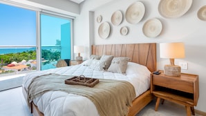 A modern bedroom with a large bed, a wooden headboard, and a view of the ocean. - Stylish Ocean-view Condo With Pool, Gym, Private Balcony, AC, & Wifi (Nuevo Nayarit)