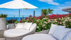 A wicker lounge chair with white cushions, a white umbrella, and a view of the sea. - Stylish Ocean-view Condo With Pool, Gym, Private Balcony, AC, & Wifi (Nuevo Nayarit)