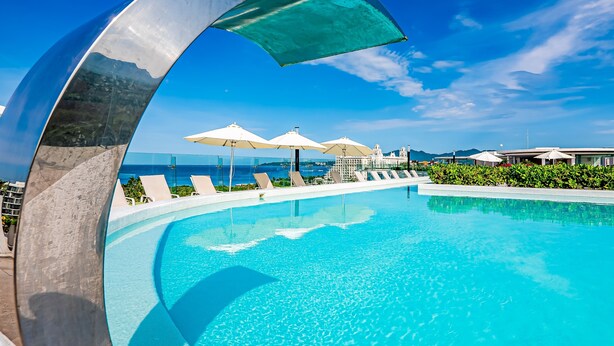 A clear blue pool with lounge chairs and umbrellas, surrounded by greenery and overlooking a scenic view. - Stylish Ocean-view Condo With Pool, Gym, Private Balcony, AC, & Wifi (Nuevo Nayarit)