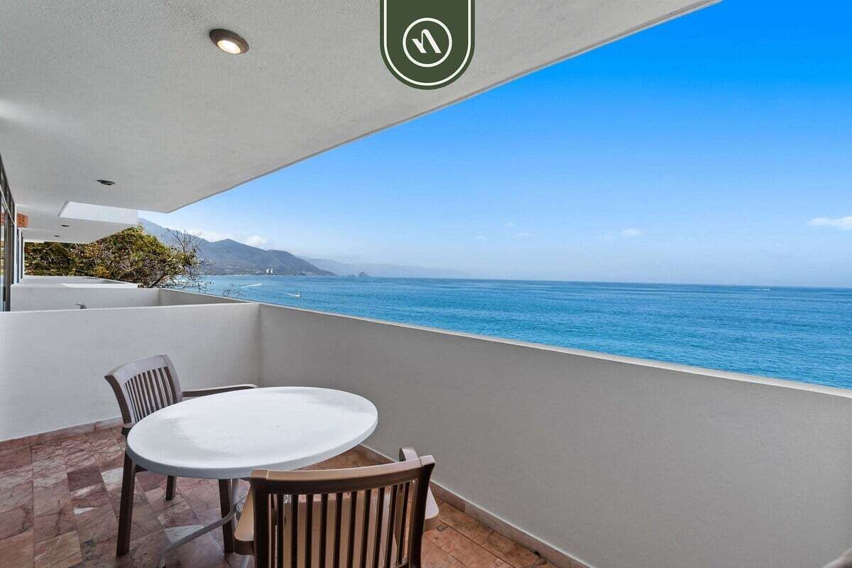 Een balkon met een witte tafel en stoelen, met uitzicht op een heldere blauwe zee en verre bergen.