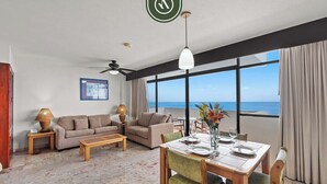 A spacious living room with a large window offering a view of the sea, a dining area with a table set for two, and a sofa set. - Costa Sur Suite 2 - Oceanview 1 BR Condo - Pool - Cleaning Included (Puerto Vallarta)