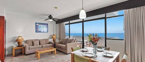 Amplia sala de estar con un ventanal que ofrece vista al mar, un comedor con una mesa preparada para dos personas y un conjunto de sofás.