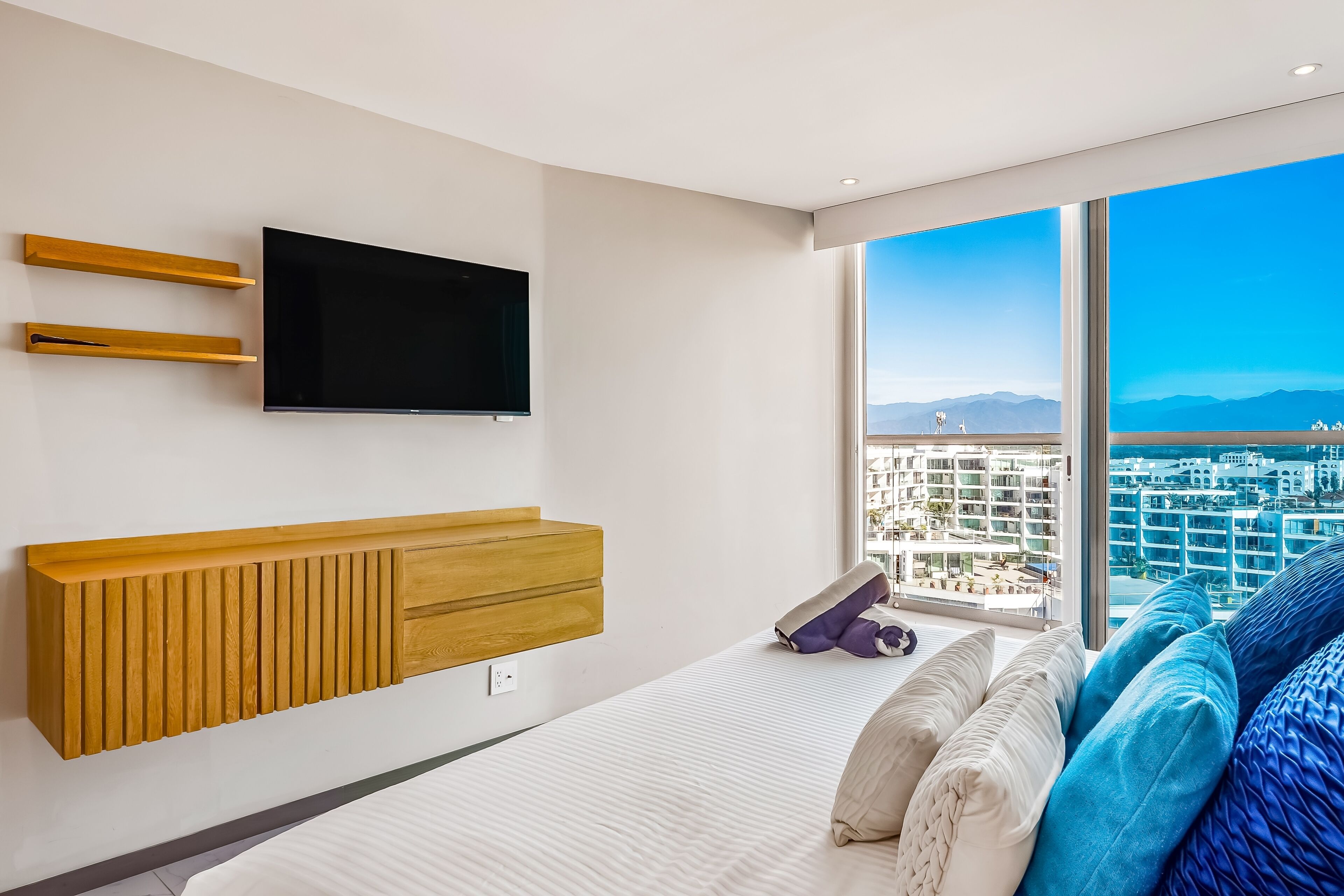 Een moderne slaapkamer met een groot raam, een flatscreen televisie, houten opbergruimte en een bed met kussens.
