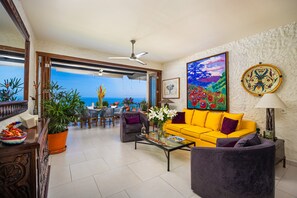 Ein Wohnzimmer mit einem gelben Sofa, einem violetten Sessel, einem Couchtisch und Blick auf das Meer.
