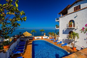 Ein Poolbereich mit blauen Liegestühlen, einem Reetdachschirm und Blick auf das Meer.