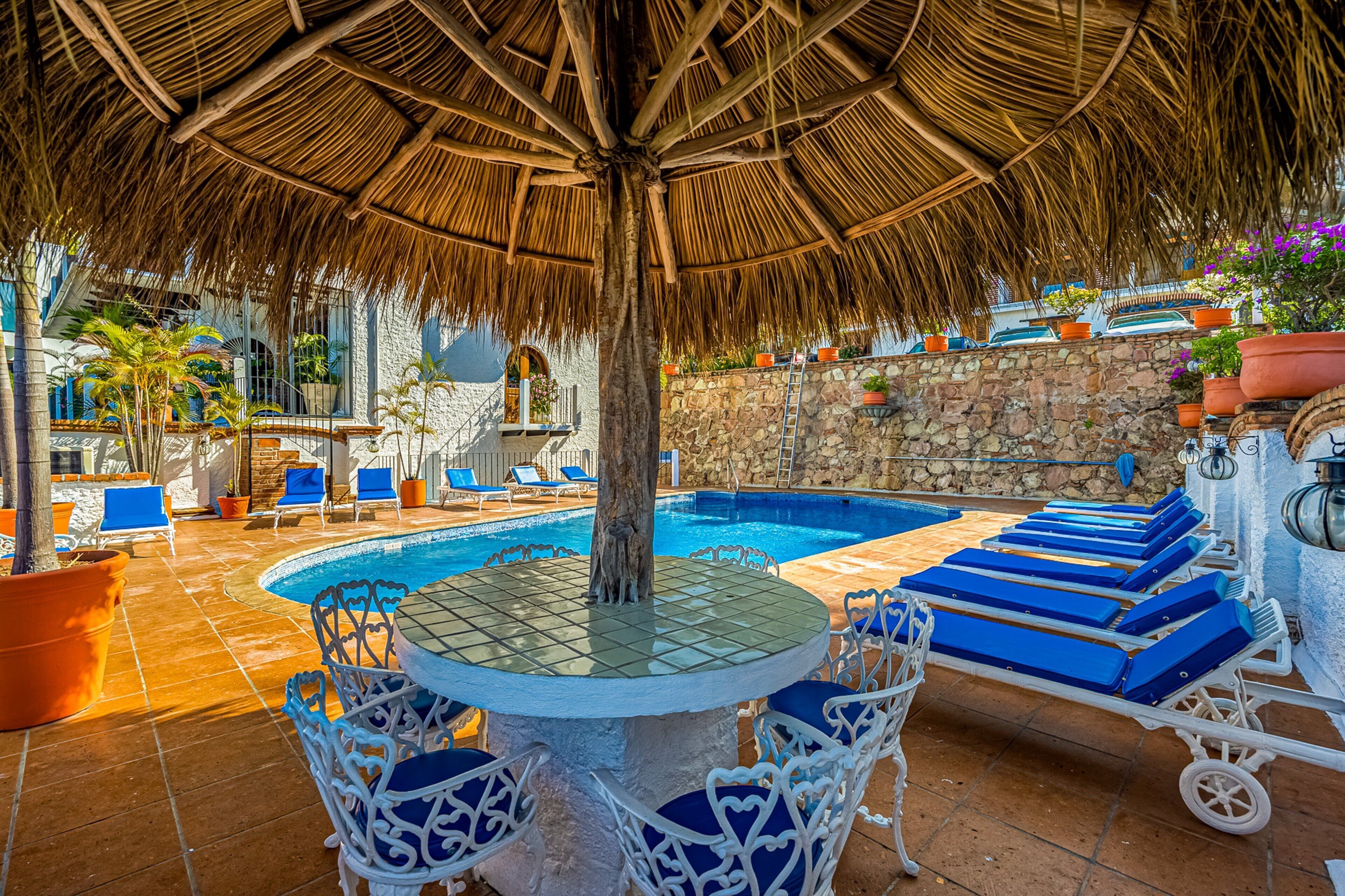 A pool area with a thatched roof, lounge chairs, and a table with chairs.
