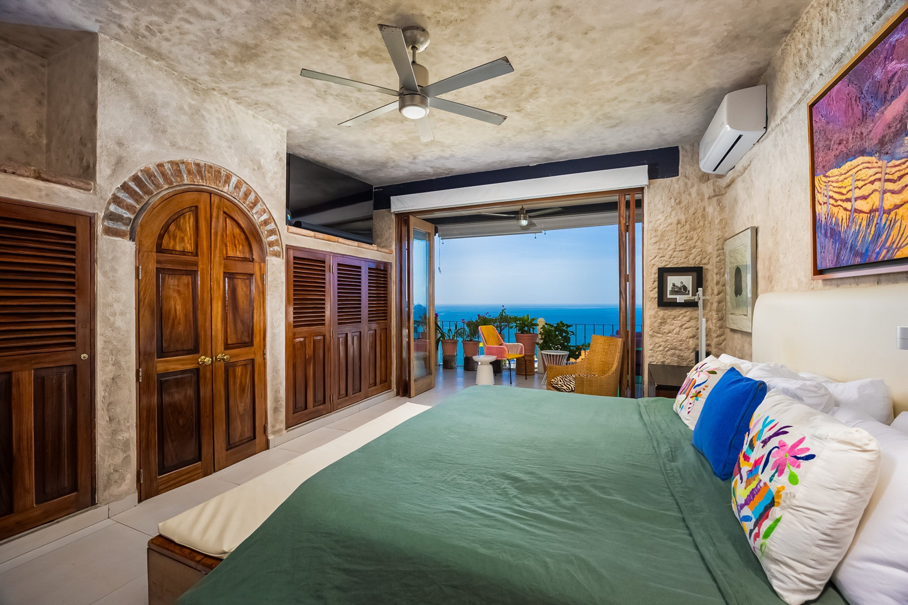 A bedroom with a large bed, a ceiling fan, a view of the ocean, and a balcony with outdoor seating.
