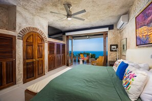 Ein Schlafzimmer mit einem großen Bett, einem Deckenventilator, Meerblick und einem Balkon mit Sitzgelegenheit.