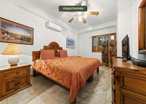 Ein Schlafzimmer mit einem Bett, einem Nachttisch, einer Lampe, einem Deckenventilator, einer Klimaanlage, einem Fernseher und einem Bild an der Wand.